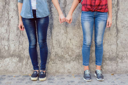 Help lesbians people comfort console concept. Close up photo of two cute lovely sweet teen age girls holding little fingers wearing stylish jeans isolated on concrete gray wall backgroundの写真素材