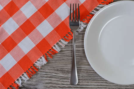 Round white empty plate, fork and checkered tablecloth on wooden table. Top view photoの写真素材