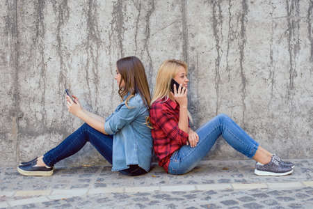 Addiction concept. Two charming cute beautiful sweet charming brunette and blonde girls using modern smartphones sitting back to back spending time separately isolated on concrete gray wall backgroundの写真素材