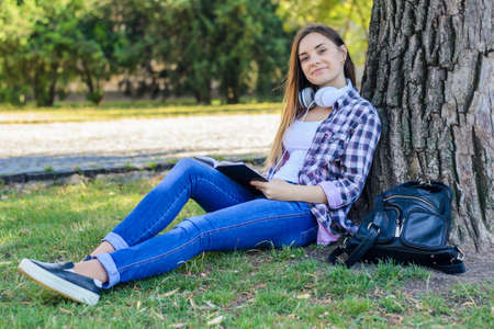 Happy smiling young girl in casual clothes with headphones on neck sitting on grass and reading bookの写真素材