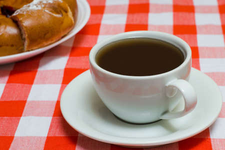 Cup of coffee on checkered red tablecloth with cake on the background. Close up photoの写真素材
