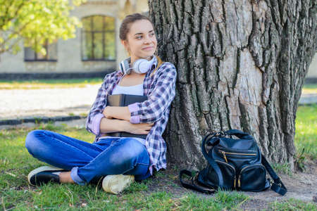 Smiling happy young woman in checkered shirt and jeans sitting on grass with book in hands and dreamingの写真素材