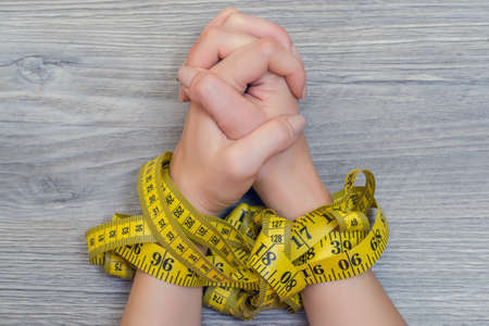 Concept of unhealthy food habbits. Close up photo of woman's hands tied with tape measure over wooden backgroundの写真素材