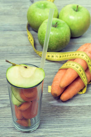 Concept of healthy nutrition and dieting. Photo of glass with straw full of slices of fruits with green apples and carrots with tape measure on backgroundの写真素材