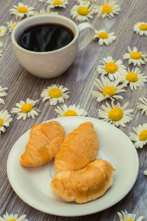 Vertical photo of romantic freah and tasty breakfast. Croissant and cup of coffee on wooden table with chamomilesの写真素材