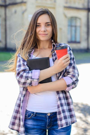 Pretty young smiling cheerful female student in casual clothes holding book and cup of coffee against campusの写真素材