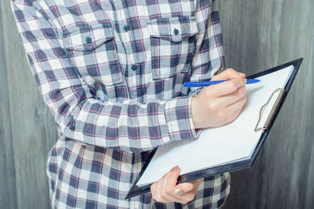 Cropped photo of young woman in checkered shirt holding clipboard and writing something on itの写真素材