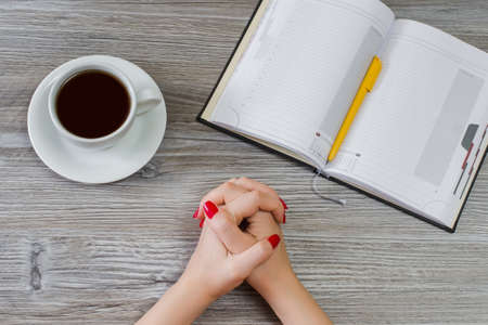 Concept of making a choice between education and having a rest. Top view photo of crossed woman's hands, cup of coffee, opened notepad with pen on wooden tableの写真素材