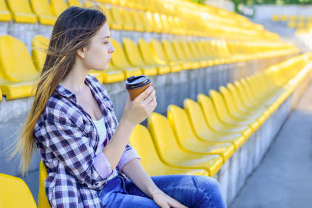 Peaceful young woman in checkered shirt with cup of coffee watching game at the stadiumの写真素材