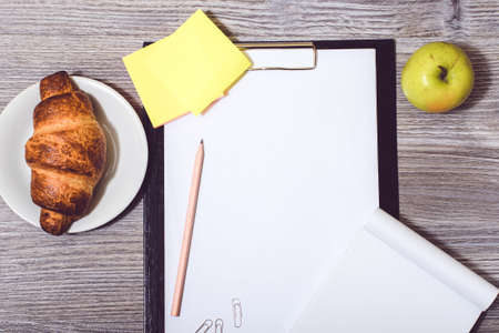 Overhead view of office accessorise: clipboard, tasty, croissant, apple, notepadの写真素材