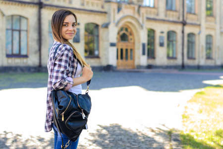 It is the first day at university for young but confident and smiling female student in checkered shirt with backpack on her shoulders. She is smiling at the camera. Entrance to the university is depicted on the backgroundの写真素材
