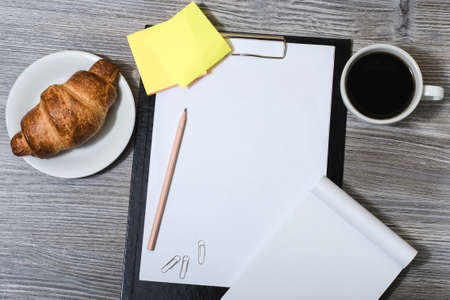 Office accessorise on wooden gray table: clipboard, cup of coffee, fresh croissantの写真素材