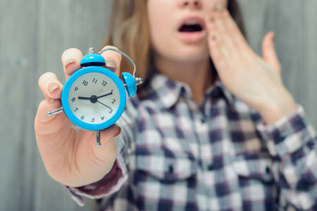 Closeup photo of alarm clock in hand and yawning woman on backgroundの写真素材