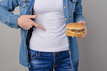 Concept of unhealthy eating and overeating. Woman has problems with her stomach. Woman holding big greasy burger and pointing on her stomach; cropped photo, close-upの写真素材