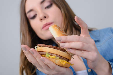Concept of unhealthy nutrition. Close up photo of curious woman looking inside of big fatty hamburgerの写真素材