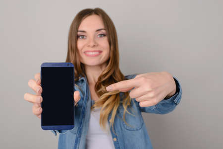 Smiling pretty woman showing black empty screen of her smartphone and pointing on it with her...
