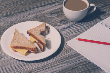 Break at work. Bitten ham and cheese sandwich on a white plate, cup of hot white coffe, open notebook with a pencil on it on wooden table. Background, vintage effect の写真素材