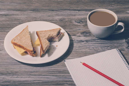 Close up of bitten cheese sandwich, cup of hot coffee, open notebook with pencil on wooden background. の写真素材