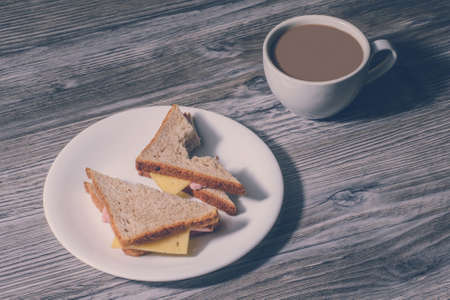 Lunch at work. Close up of bitten cheese sandwich on a white round plate and cup og hot coffeeの写真素材