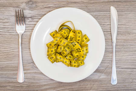 Concept of unhealthy nutrition. Plate with tape measure, fork and knife on wooden background. Overhead view photo.の写真素材