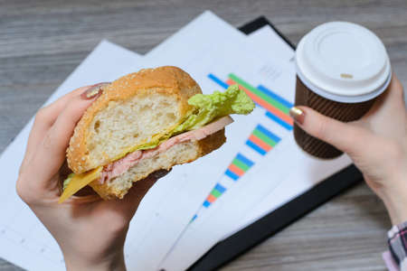 Break at work. Hungry worker eating cheeseburger and drinking coffeeの写真素材