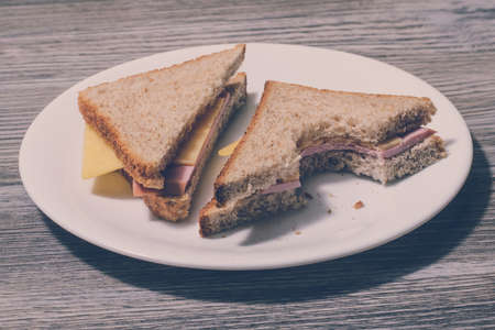 Tasty bitten ham sandwich on a white plate, close up, wooden background, horizontal view, vintage effectの写真素材