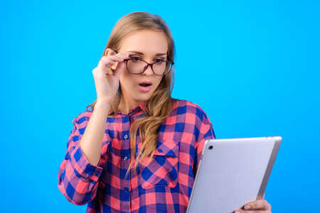 Close up portrait of astonished pretty cute charming with long blonde hair shocked teenager touching glasses looking at tablet in hands wearing casual outfit isolated on blue backgroundの写真素材