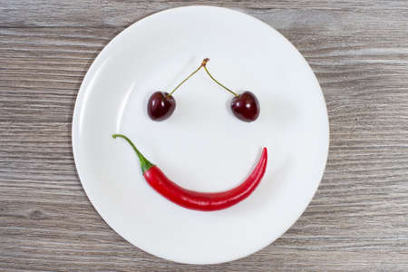 Happy smiling face made of cherry and chili pepper on white plate. Top view, wooden tableの写真素材