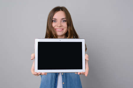 Attractive woman with toothy smile showing digital tablet with black blank touchscreen. The woman is isolated on grey backgroundの写真素材