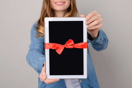 Woman showing digital tablet with red ribbon gift isolated on grey backgroundの写真素材