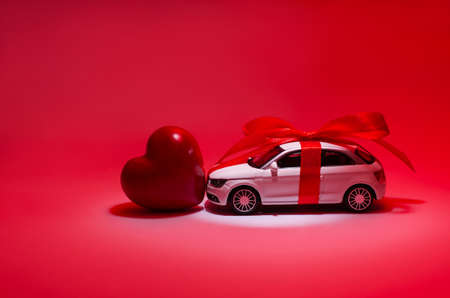 Profile side close up view photo of beautiful white toy modern fashionable trendy car with red ribbon and bow red heart in center in spotlight searchlight isolated on red background copy-spaceの写真素材