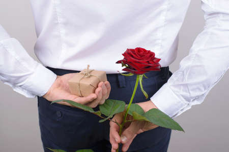 Back behind rear view photo of happy romantic charming handsome gentleman hiding beautiful rose and small package isolated grey backgroundの写真素材