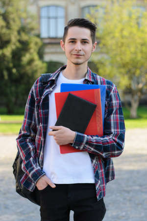 Vertical full length body size photo of handsome nice cheerful excited millenial holding stack of book in hand standing in front of buildingの写真素材