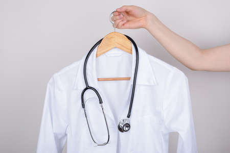 Job vacancy concept. Cropped close up photo of hand holding white doctor coat on hanger isolated over grey backgroundの写真素材