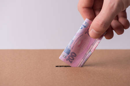 Medical insurance concept. Cropped closeup photo of man human male hand putting ukrainian money in hole in cardboard boxの写真素材