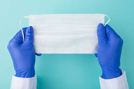 Doctors staying safe during Coronavirus outbreak concept. Top above overhead view photo of doctor in gloves holding a white surgical mask isolated on turquoise backgroundの写真素材