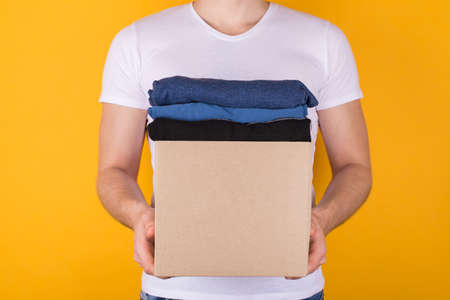 Donation concept. Cropped front view photo of man holding cardboard box full of clothes isolated on yellow backgroundの写真素材