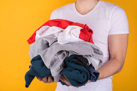 Laundry concept. Cropped half turned photo of man holding lots of clothes isolated on yellow...