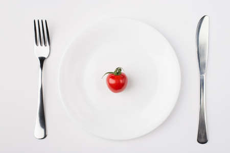Eating a low calorie diet concept. Top above overhead flat-lay view photo of a plate with a single tomato with fork and knife aside isolated on white backgroundの写真素材