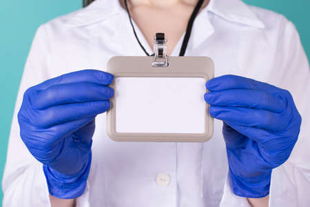 Close-up cropped photo of female doctor in white coat uniform gloves holding a blank mock up nametag in front isolated on blue turquoise  backgroundの写真素材