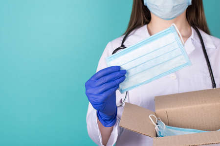 Humanitarian aid help support concept. Close-up cropped photo of female doctor in gloves holding a box of masks and one in hand isolated on blue teal turquoise backgroundの写真素材