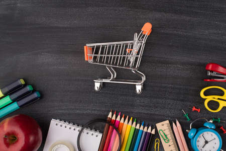 Back to school stationery shopping advert concept. Top above overhead view photo of small cart in center apple and colorful stationery isolated on blackboard with copyspaceの写真素材