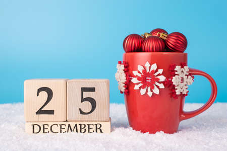 25th december concept. Close up photo of red stylish mug full of baubles and wooden calendar standing in snowの写真素材