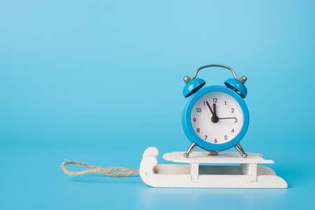 Five minutes before new year concept. Close up photo of small toy miniature clock showing time to midnight standing on sledges isolated blue bright color backgroundの写真素材
