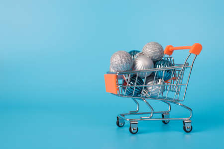 Christmas shopping concept. Close up photo of miniature push cart full of vivid baubles isolated on blue background with copyspaceの写真素材