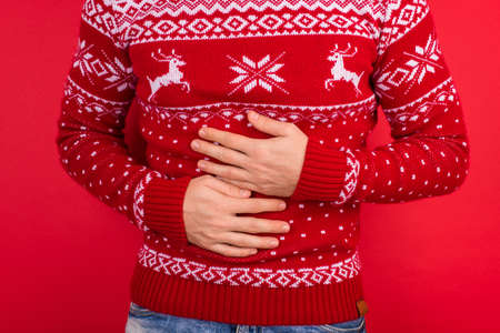 Bellyache concept. Close up photo of men in red winter sweater with deers holding his stomach isolated on red backgroundの写真素材