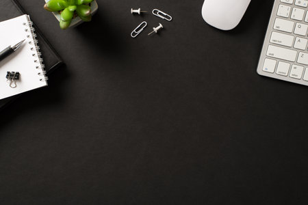 Flatlay above overhead view photo of white office items computer keyboard mouse pins paper clips green succulent and several books and spiral notepads isolated on black backdropの写真素材