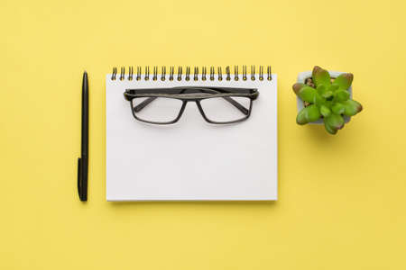 Photo overhead of notebook spectacles plant and black pen isolated on the yellow backgroundの写真素材