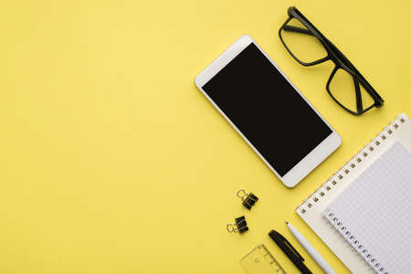 Photo overhead of notebook glasses paperclips phone pen and straightedge isolated on the yellow...