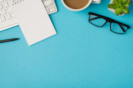 Top view photo of desktop with keyboard notepad pen glasses plant and cup of coffee on isolated blue background with blank spaceの写真素材